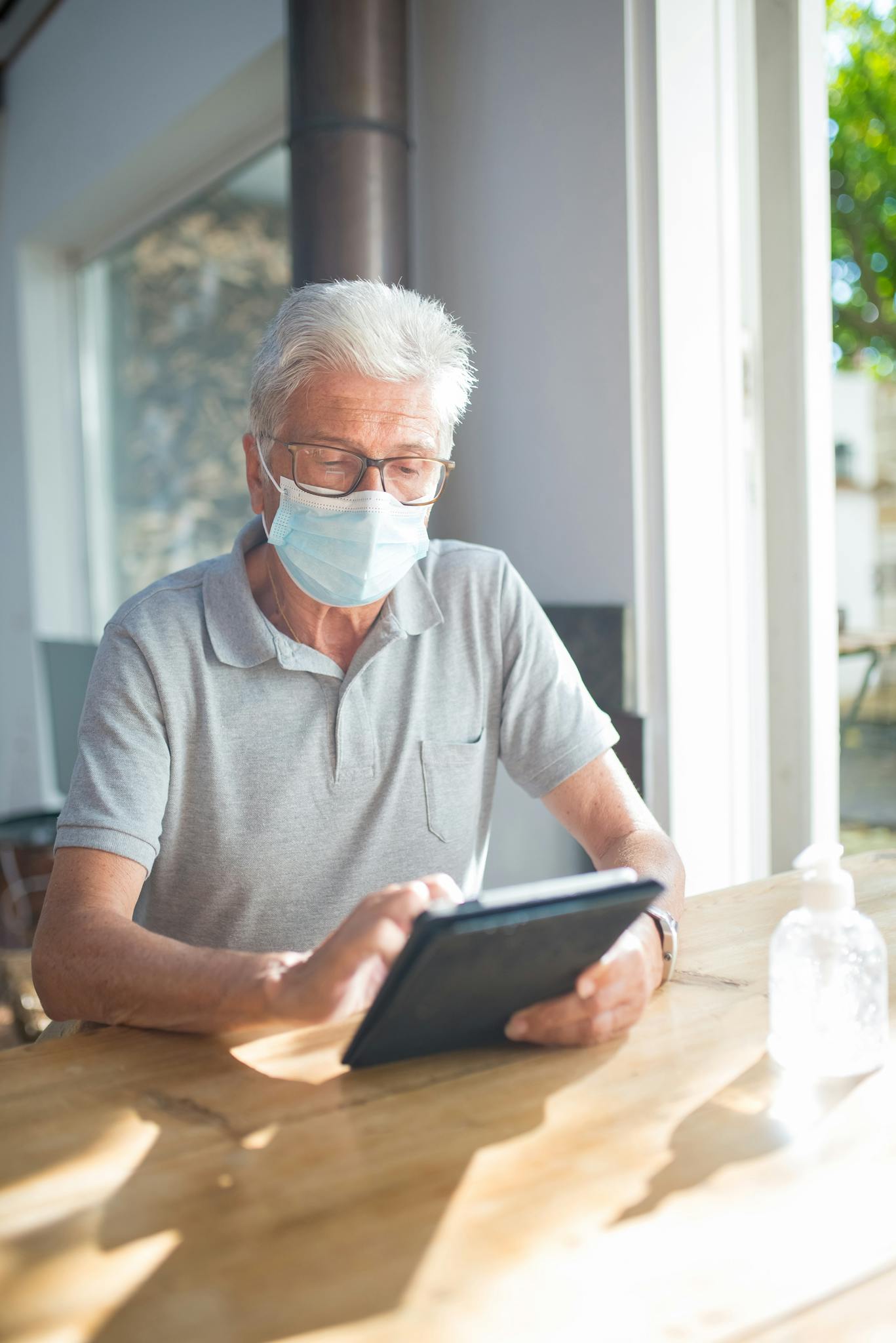 Elderly man wearing face mask using tablet indoors. Sunlit modern setting, health protection focus.